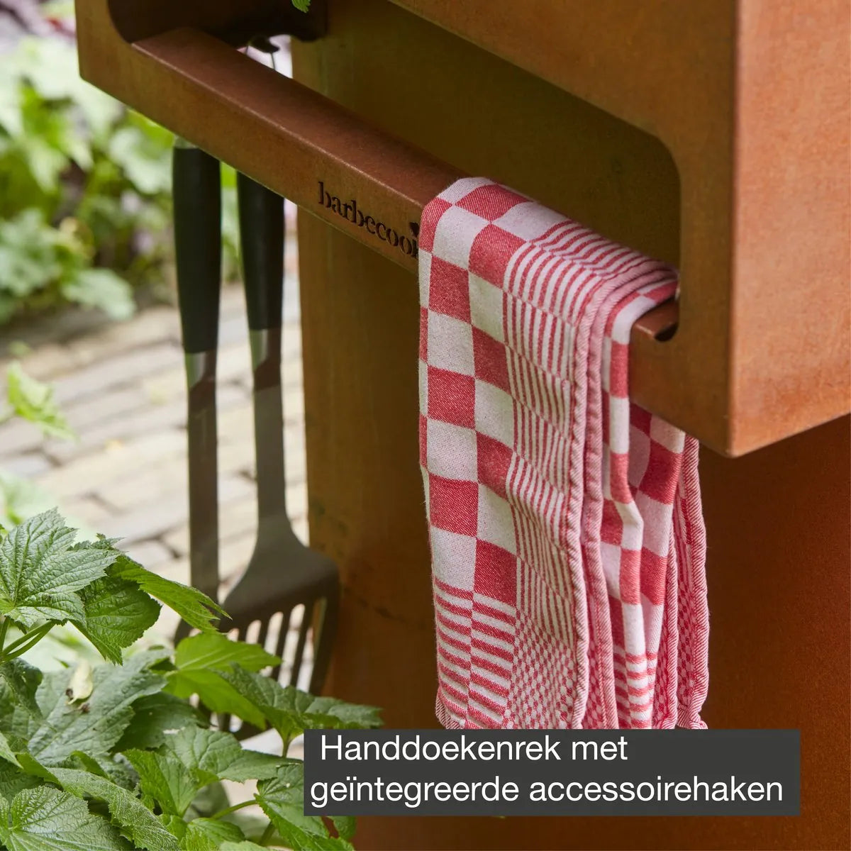 A red and white checkered kitchen towel hangs from the handle of a corten steel barbecook nestor island wood plancha grill