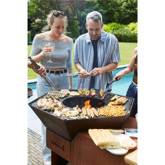 Black hexagonal corten steel Barbecook Nestor Island grill with central fire pit cooking meats vegetables and chickpeas