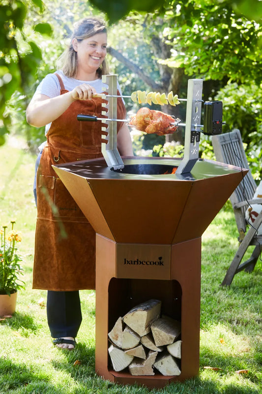 Rust-colored metal barbecue with built-in wood storage and rotisserie attachment from barbecook for nestor world