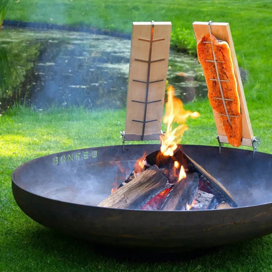 Black metal bonfeu fire pit with wooden racks holding grilled fish over flames for smoking meat and fish