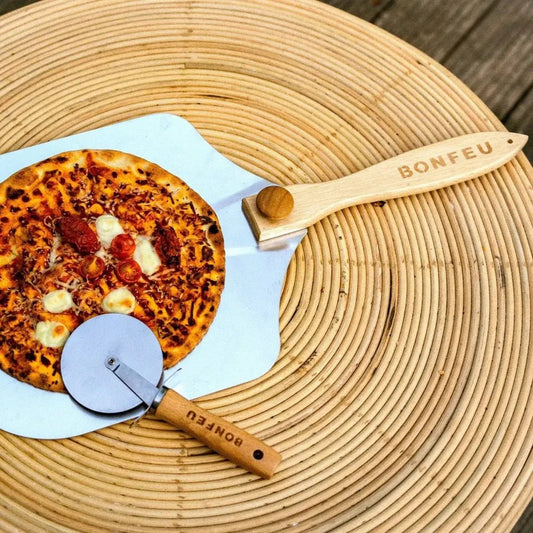 Wooden-handled bonfeu pizza cutter with metal blade resting beside a freshly baked pizza on white peel