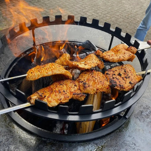 Black metal bonfeu skewers ring fire pit with flame and skewered grilled chicken pieces inside for outdoor cooking