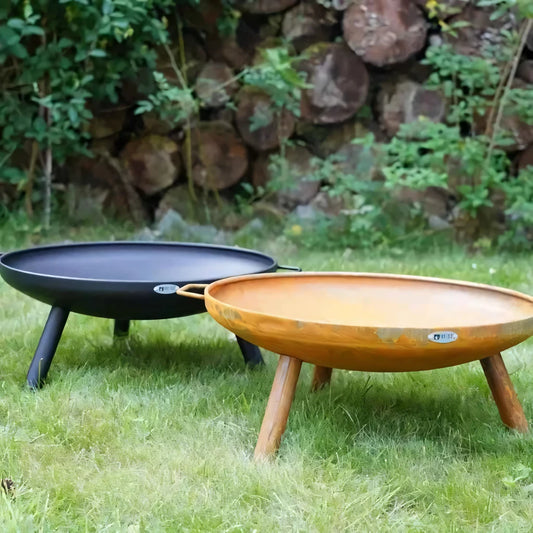 Two round three-legged heat fire bowl classic feet and handle 60 cm fire pits in black and rust on grass with log wall background