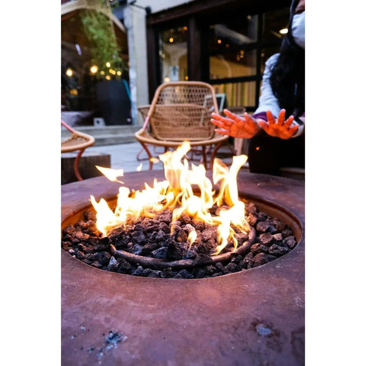 Rust-colored metal fire pit with flickering orange flames and dark volcanic rocks in a garden setting