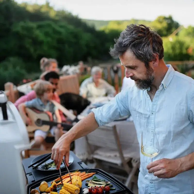 Man grilling over a small gas BBQ with friends in the background