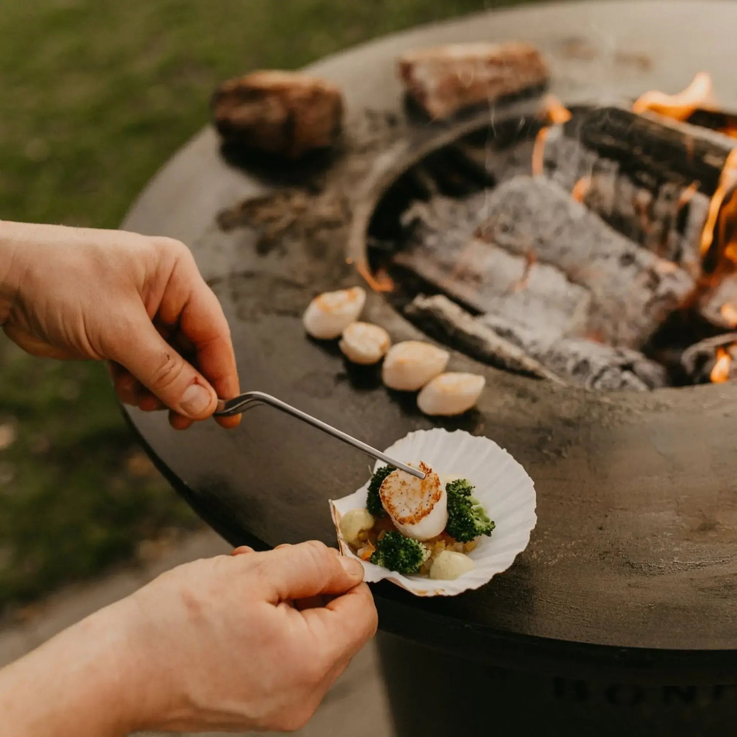 A seared scallop on broccoli and garlic served on a white shell plate beside a black bonfeu bonbiza open plancha grill fire pit