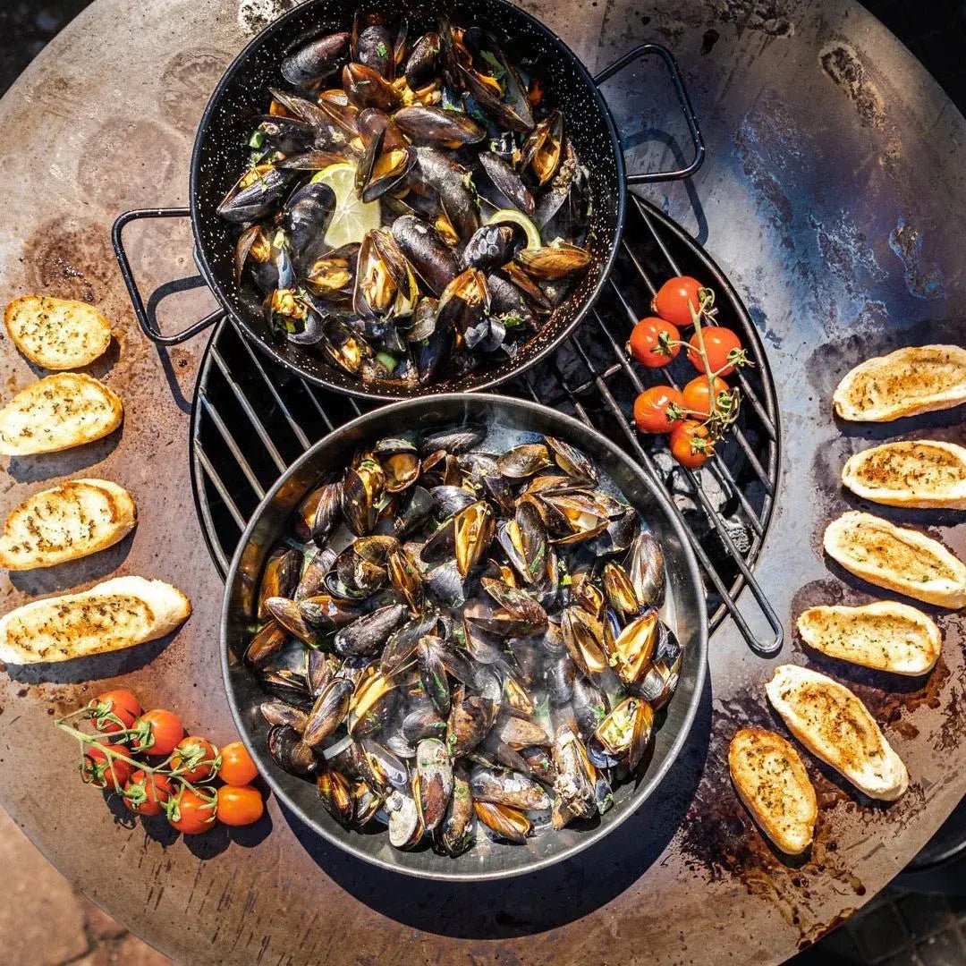 Black speckled cast iron pans filled with steaming mussels on a bonfeu bonbiza open plancha grill fire pit