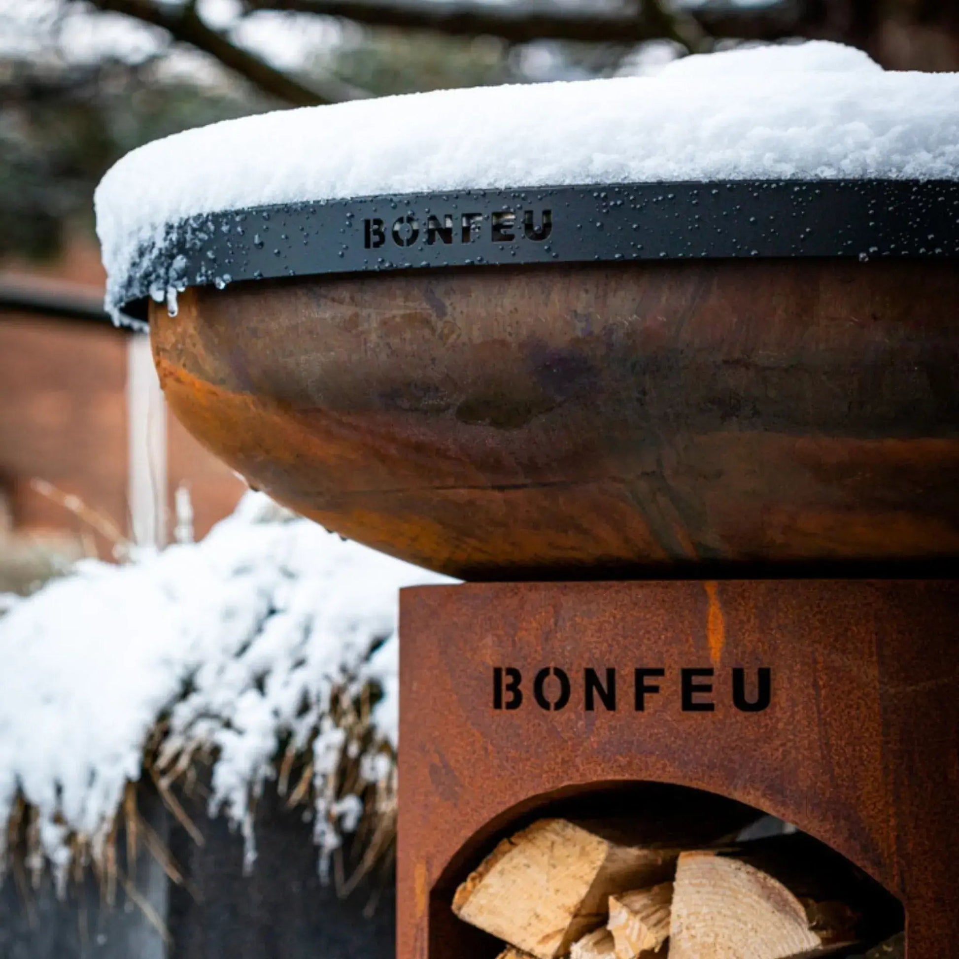 Rust-colored corten steel bonfeu bonbiza open plancha grill fire pit with snow on top and wood stacked inside