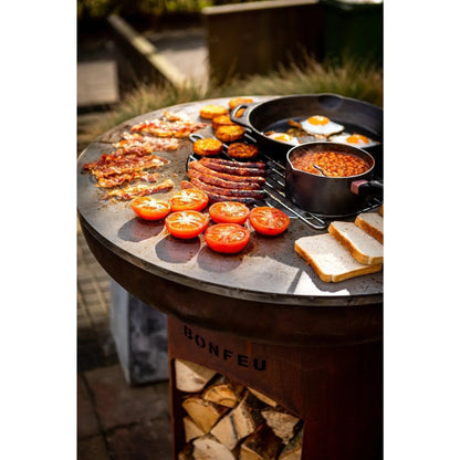 Rustic round black cast iron BonFeu BonBiza open plancha grill with stacked wood logs visible beneath the base