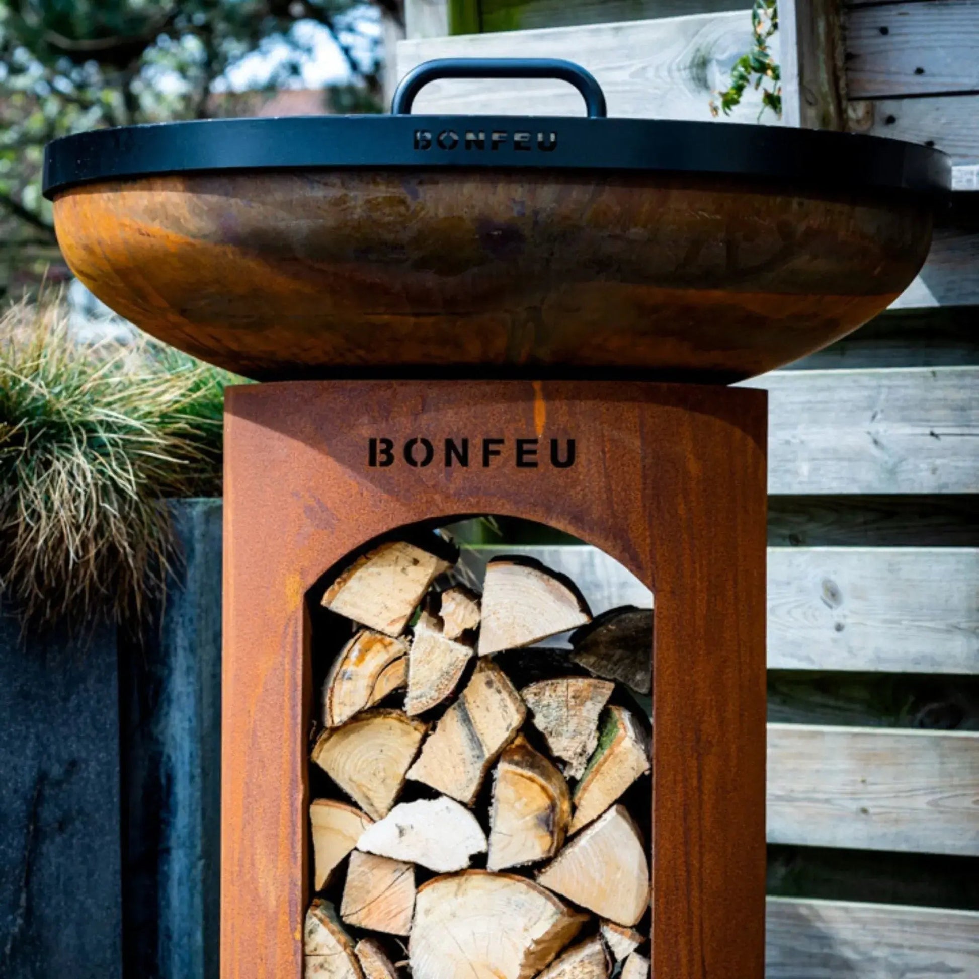 Rust-colored corten steel fire pit with black handle bonfeu branding arched opening stacked logs bonfeu bonbiza grill