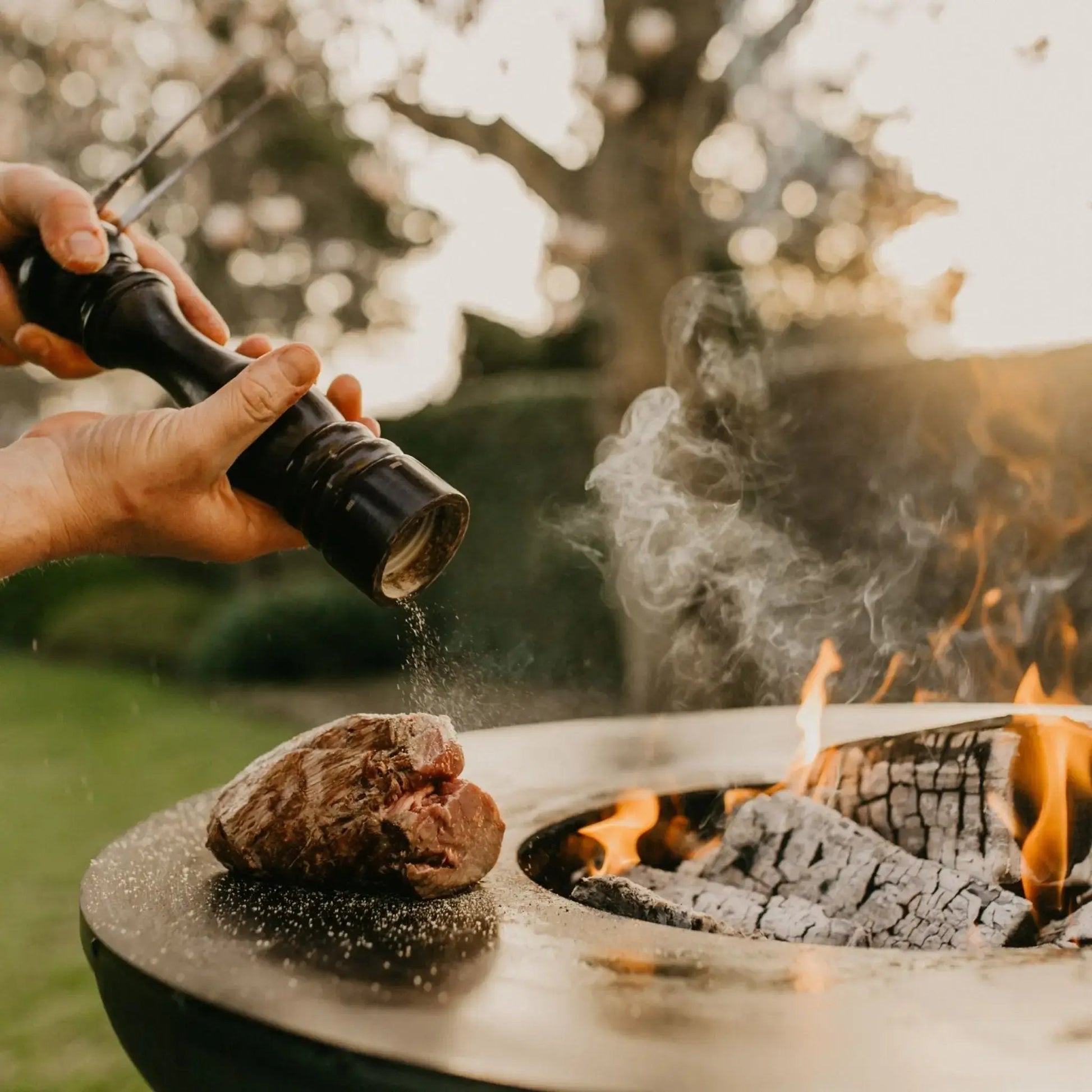 Black pepper grinder seasoning meat over bonfeu bonbiza plancha grill fire pit in black steel outdoors