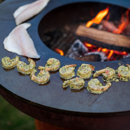 Black bonfeu bonbiza plancha grill fire pit with glowing embers cooking shrimp and fish fillets on round steel grate