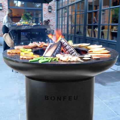 Black stone bonfeu bonbiza plancha grill fire pit with fire burning and food cooking on its surface outdoors