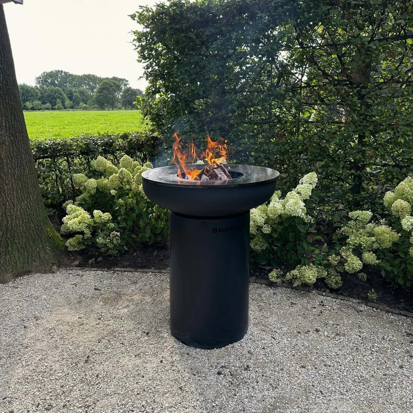 Black cylindrical bonfeu bonbiza plancha grill fire pit with flames and smoke rising from burning wood