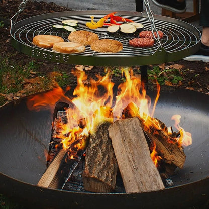 Green metal circular grill with engraved brand name hanging by chain above burning logs in bonfeu bonbowl corten steel fire pit
