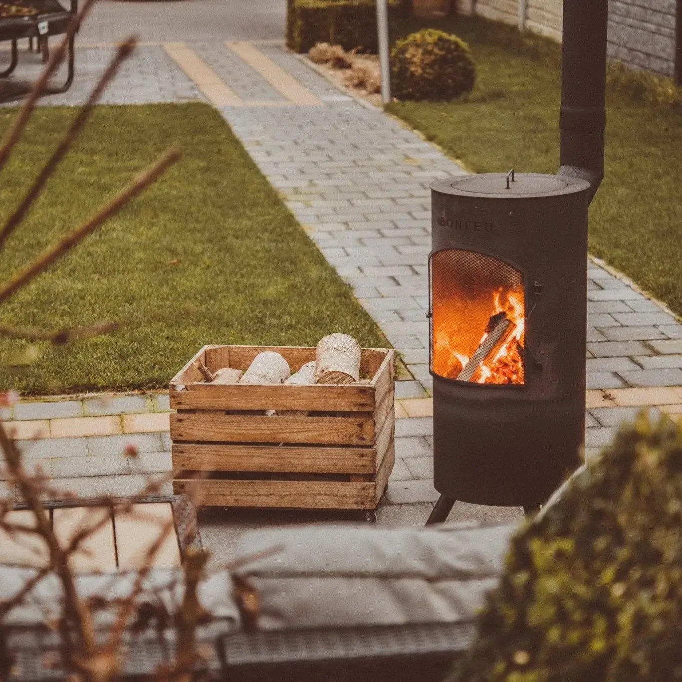 Black metal BonFeu BonTino LP outdoor wood-burning stove with mesh front and visible flames on three legs next to logs
