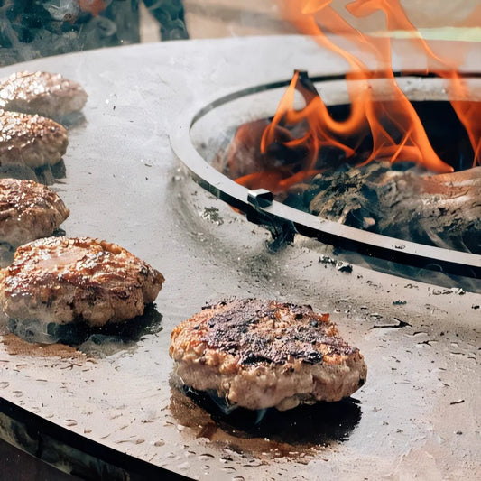 Golden-brown beef patties sizzle on a black bonfeu food bumper griddle with flames at an open fire pit