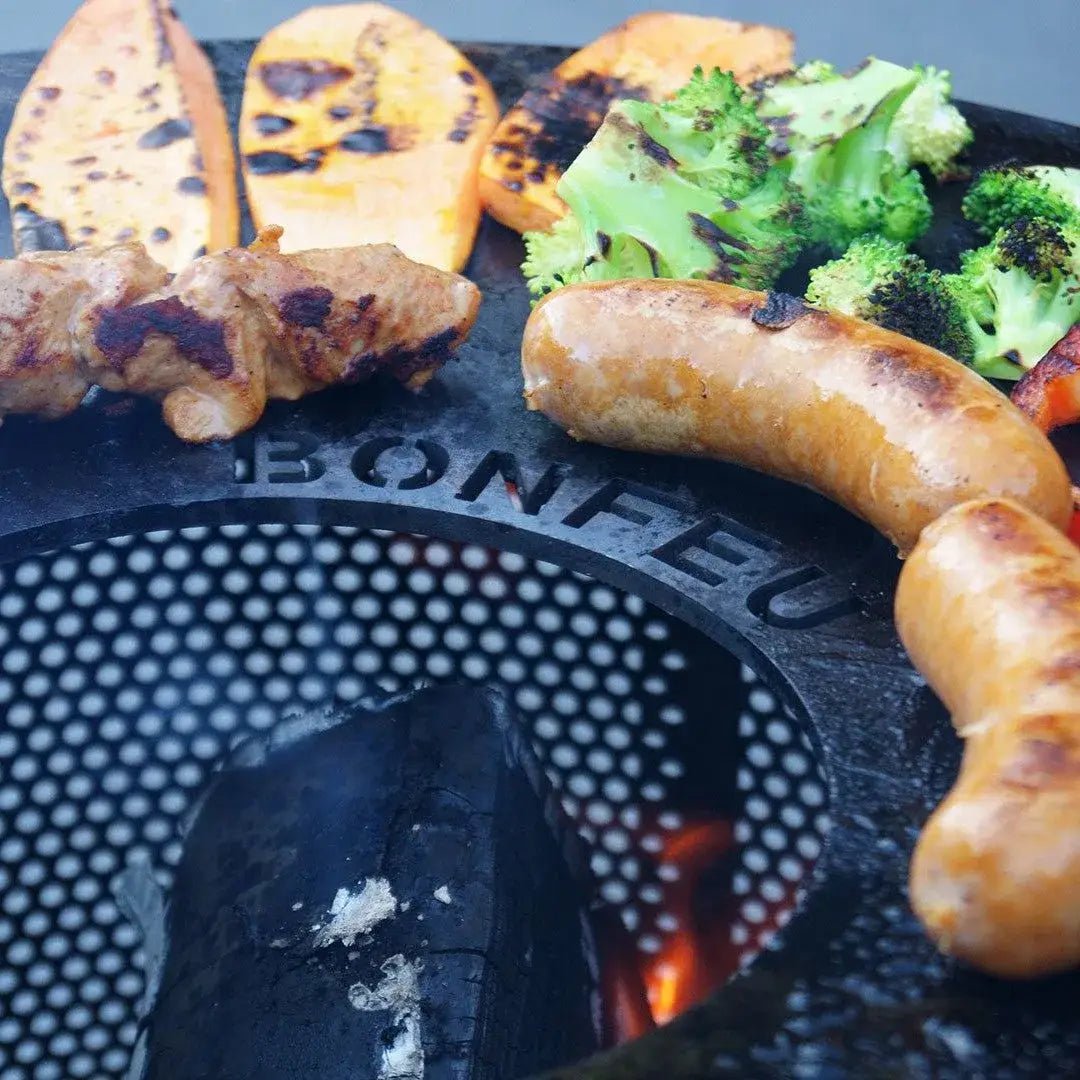Black circular bonfeu plancha grill with perforated surface cooking sausages chicken skewers roasted vegetables on embers