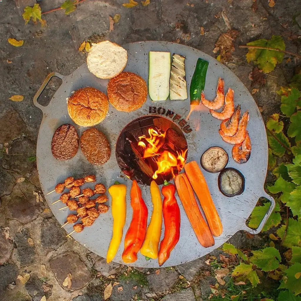 Gray circular bonfeu plancha grill with central fire pit and handles cooking burgers shrimp vegetables and skewers