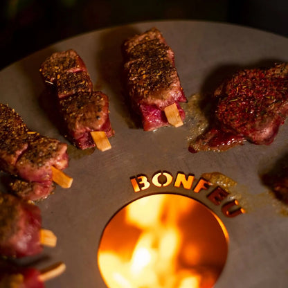 Grilled beef skewers with a seasoned charred exterior on a black circular BonFeu plancha fire pit for outdoor fireplaces