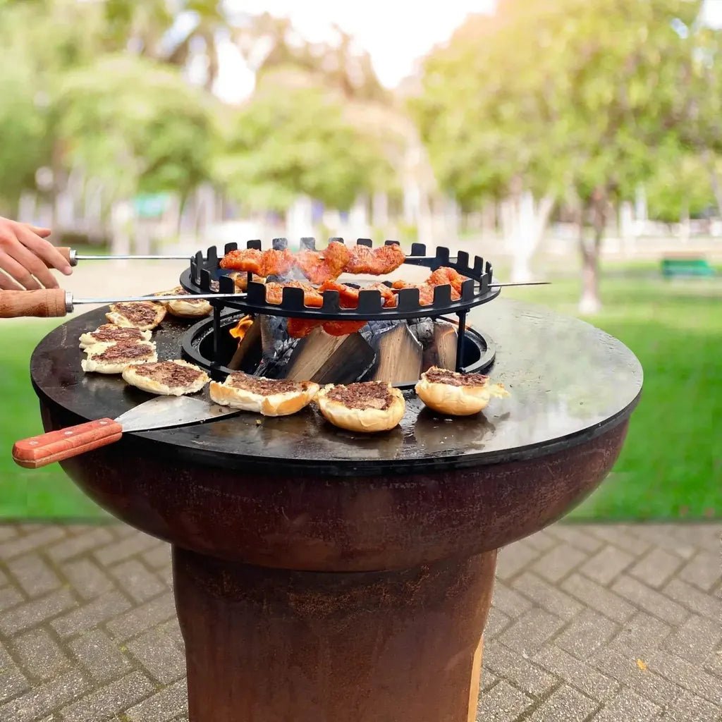Rustic round cast iron outdoor grill with black metal BonFeu skewers holding grilled meat and buns on flat surface