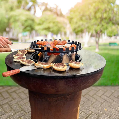 Rustic round cast iron outdoor grill with black metal BonFeu skewers holding grilled meat and buns on flat surface