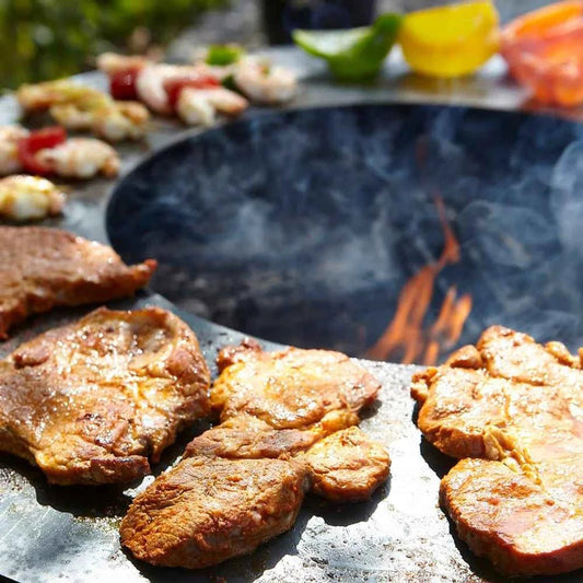 Grilled pork chops sizzling on a black stone buschbeck plancha cooking ring for 60cm fire pits with flames and smoke