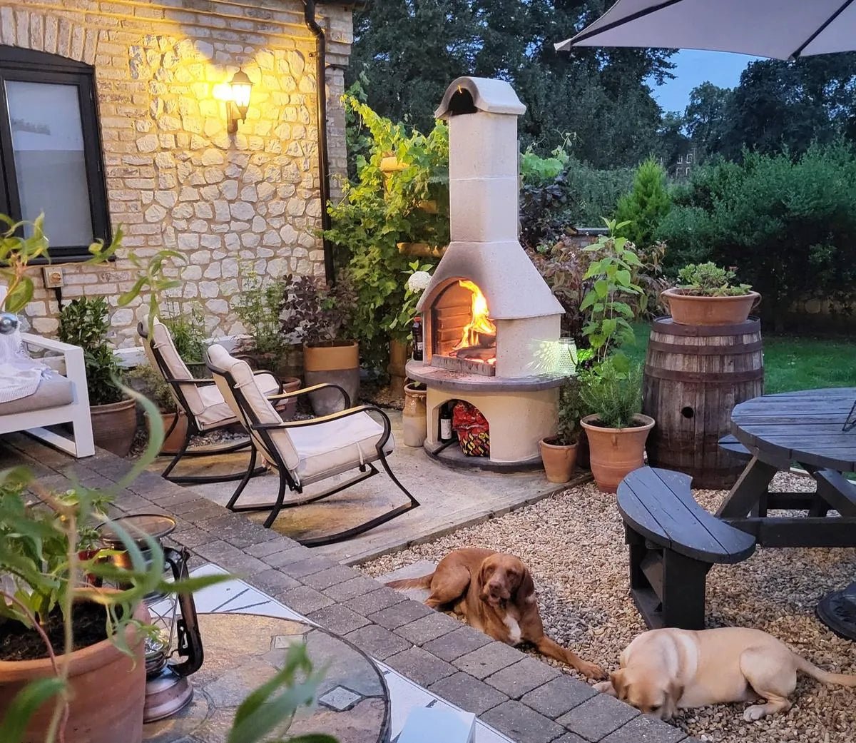 Cream-colored stone outdoor pizza oven with curved chimney and glowing fire inside from Buschbeck Verona masonry barbecue