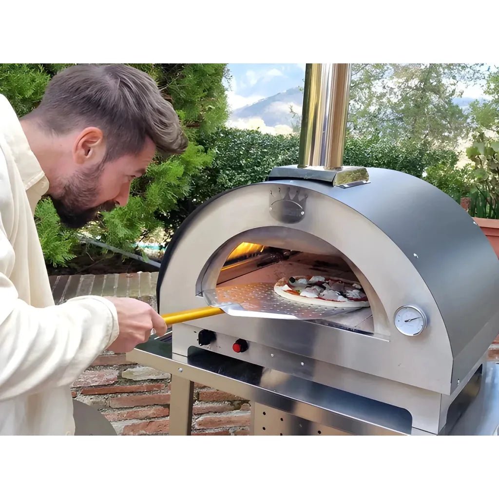A man in a cream-colored sweatshirt is using a stainless steel outdoor pizza oven, sliding a pizza in with a long yellow-handled tool. The scene is set outdoors with green trees and a brick patio in the background.