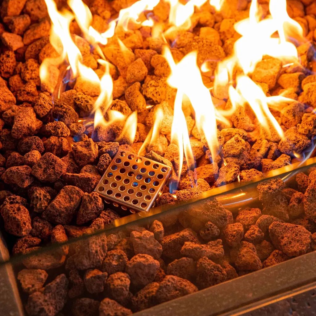 A square metallic fire spreader with circular holes on glowing lava rocks in the elementi birmingham dining fire table