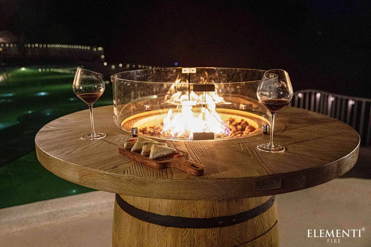 Elementi Lafite barrel bar fire table with round wooden top, built-in 13.2 kW fire pit, clear glass enclosure, and barrel base