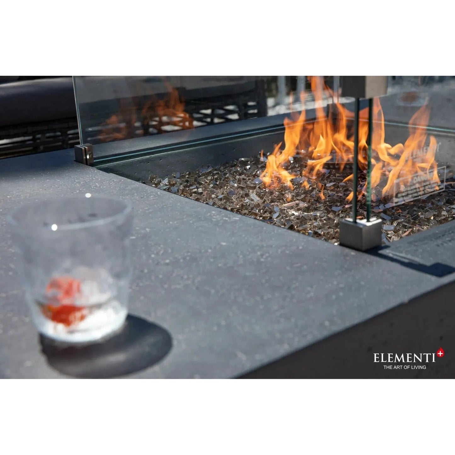 A clear glass tumbler filled with ice and a red-orange beverage on a dark gray stone countertop beside elementi plus cannes fire table 16 kw