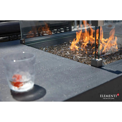 A clear glass tumbler filled with ice and a red-orange beverage on a dark gray stone countertop beside elementi plus cannes fire table 16 kw