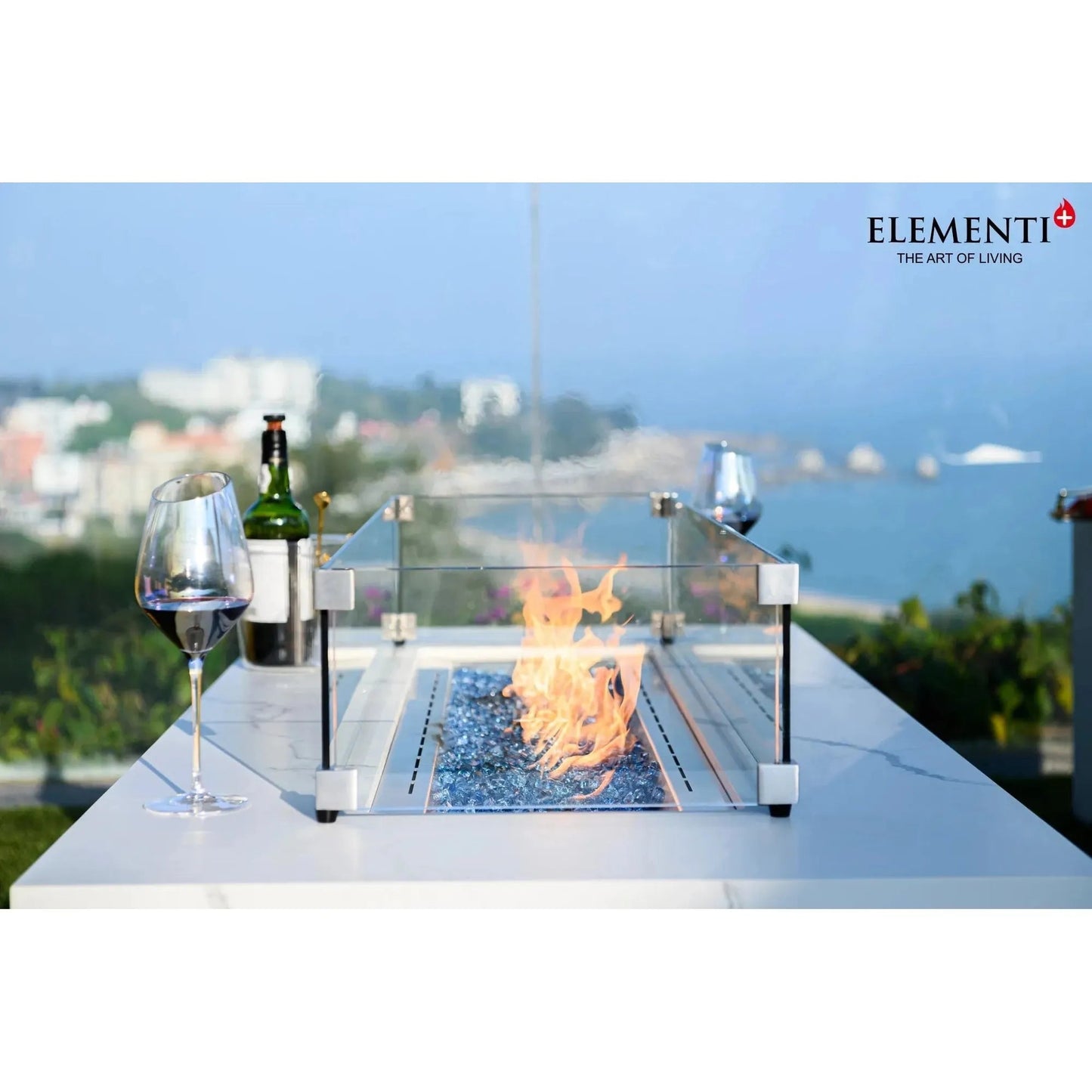 Elementi Plus Carrara fire table with silver metallic frame and glass enclosure featuring blue flameless stones and orange flame