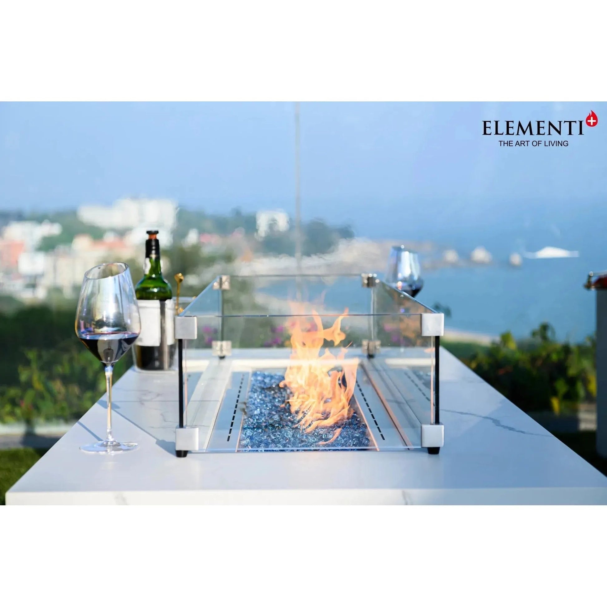 Elementi Plus Carrara fire table with silver metallic frame and glass enclosure featuring blue flameless stones and orange flame