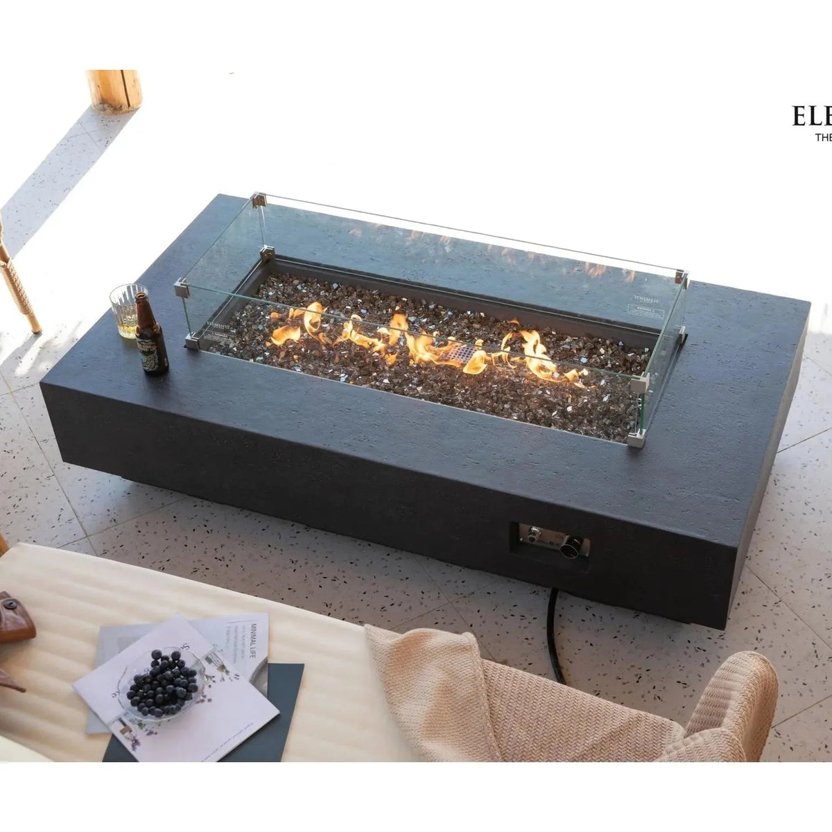Black rectangular Elementi Plus Positano fire table with glass enclosure and glowing flames over dark pebbles