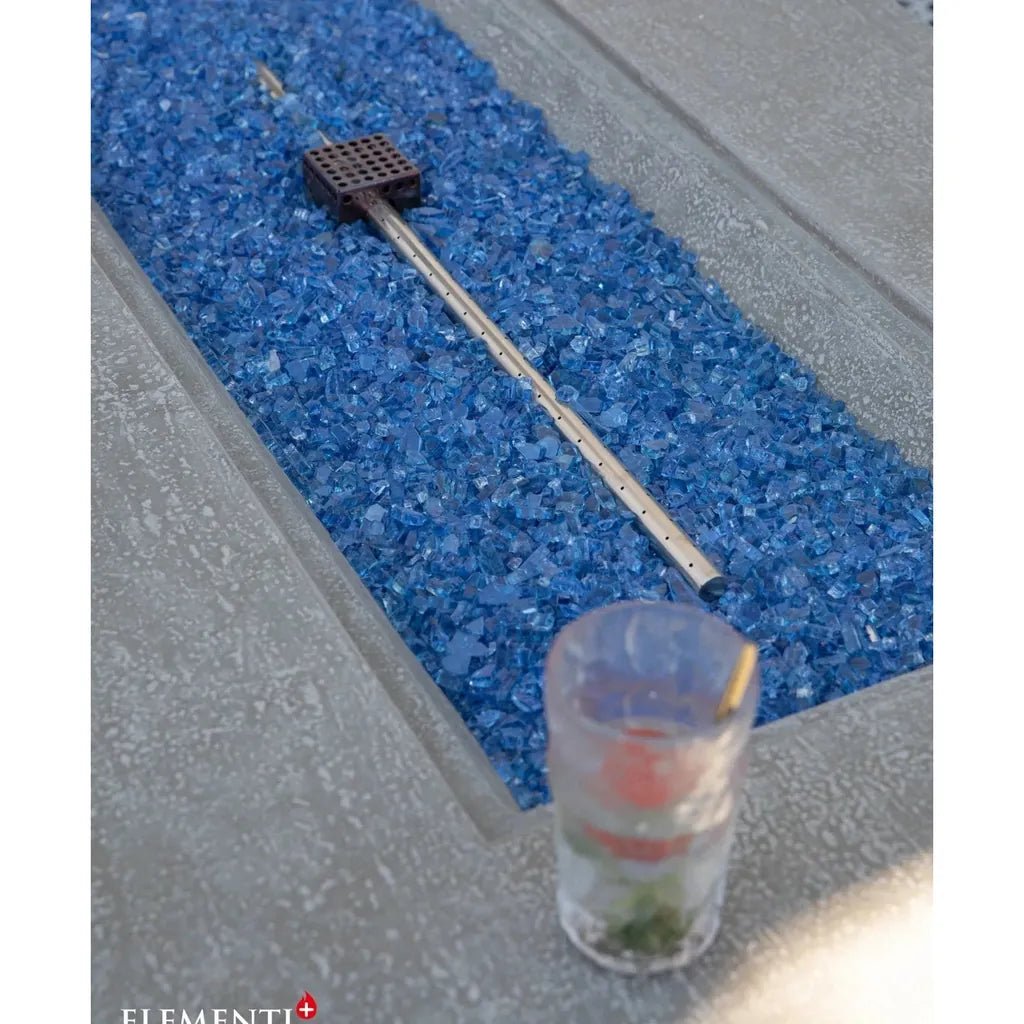 A sleek metallic fire pit tool with a black grid head resting on blue glass gravel for elementi plus riviera fire table