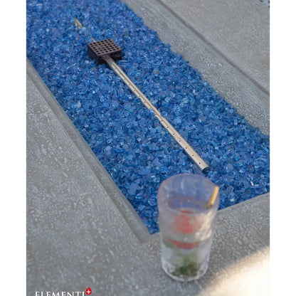 A sleek metallic fire pit tool with a black grid head resting on blue glass gravel for elementi plus riviera fire table