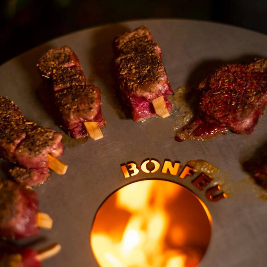Grilled beef skewers with a seasoned charred exterior on a black circular BonFeu plancha fire pit for outdoor fireplaces