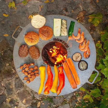 Gray circular bonfeu plancha grill with central fire pit and handles cooking burgers shrimp vegetables and skewers