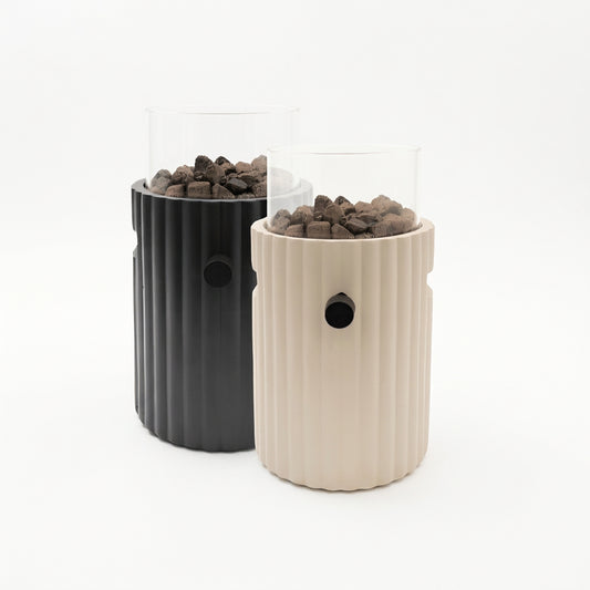 Two cylindrical Happy Cocooning Lumi Rib tabletop gas fireplaces in black and cream with ribbed textures and glass lids filled with pebbles