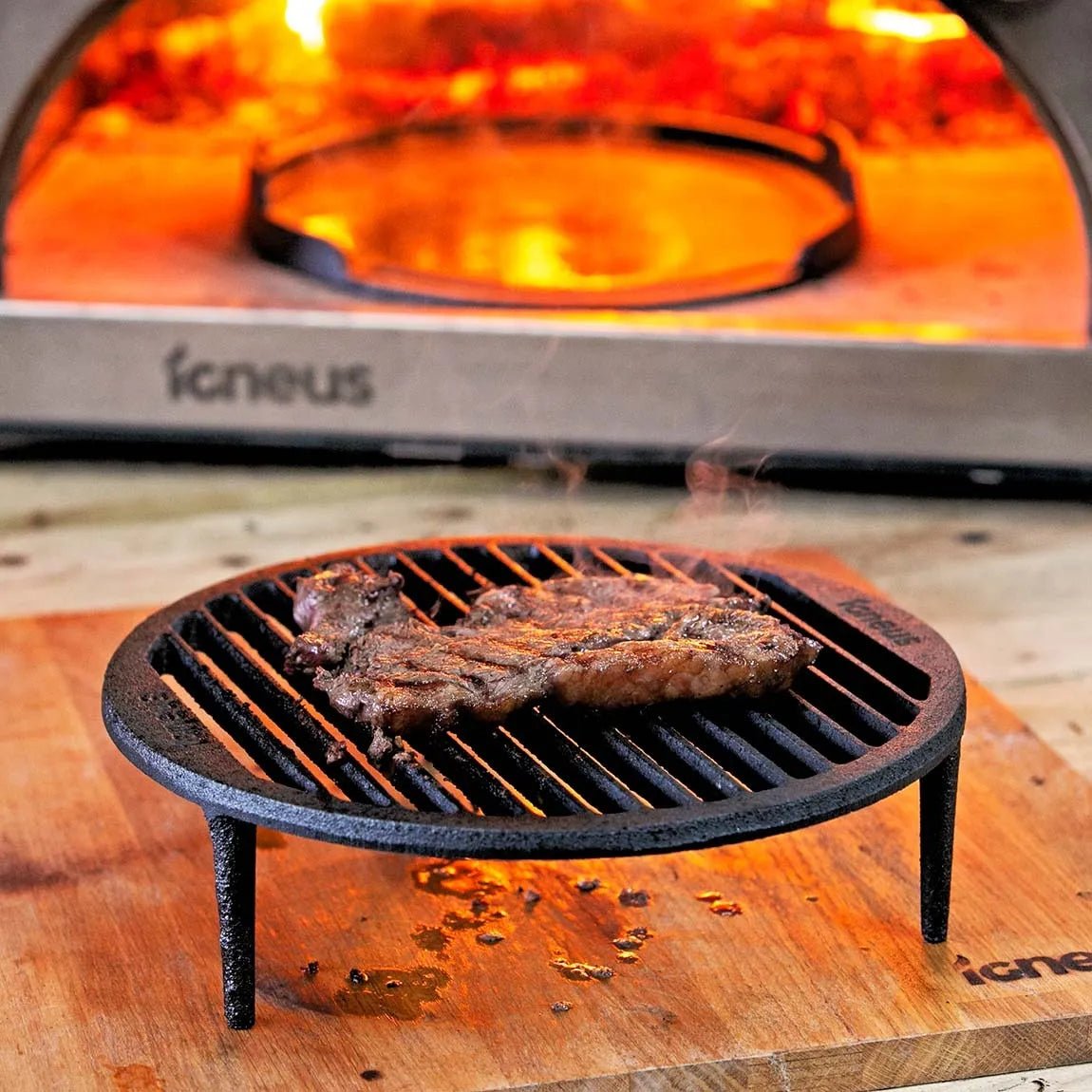 Black cast iron igneus tuscan grill grate with three legs on wood in front of orange flame pizza oven