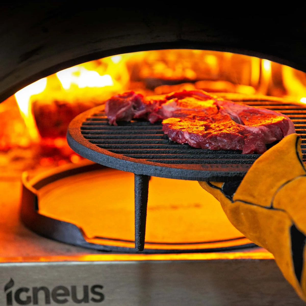 Black cast iron igneus tuscan grill grate with meat sizzling held by yellow oven mitt for pizza oven cooking