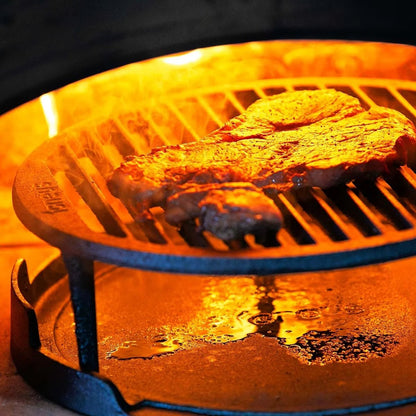 A seasoned steak sizzles on a golden-brown round cast iron grate over glowing flames on the Igneus Tuscan grill