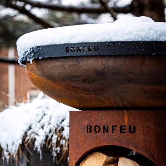 Rust-colored steel lid for BonFeu BonBiza or BonBowl fire pit with black engraved branding and snow on top