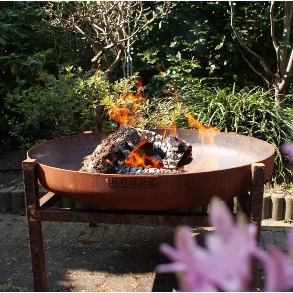 Rust-colored corten steel moodz classic fire bowl engraved with brand name outdoors among greenery and flowers