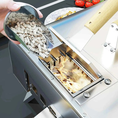 A hand holds a metallic scoop of gray wood pellets above a burning stainless steel Outsunny outdoor pizza oven