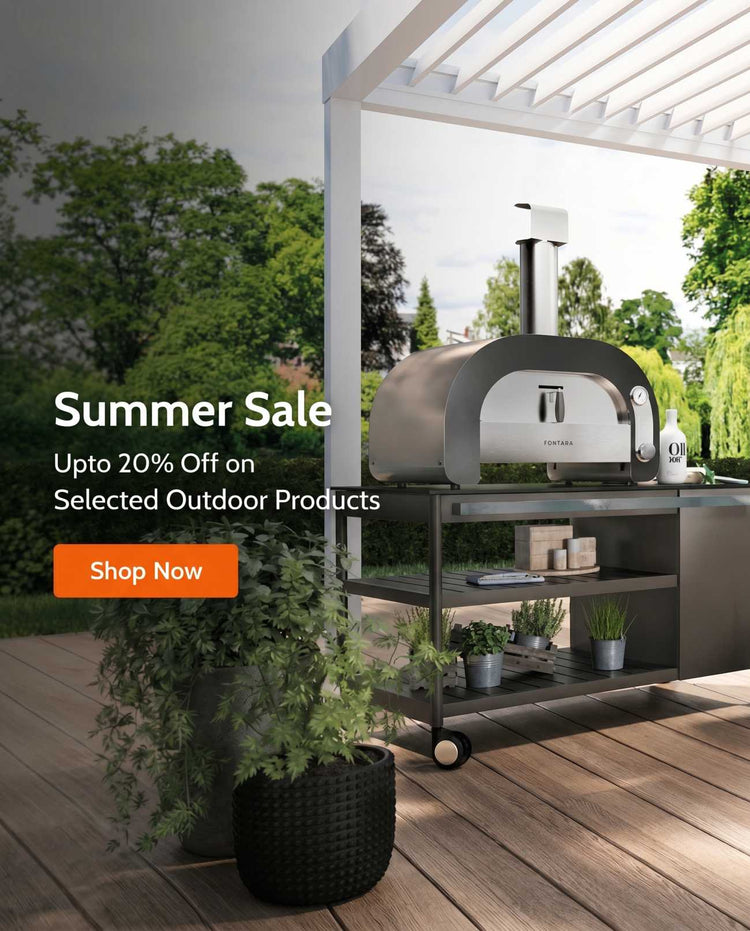 A stainless steel outdoor pizza oven on a rolling cart with shelves, set under a white pergola on a wooden deck. Green trees are visible in the background.